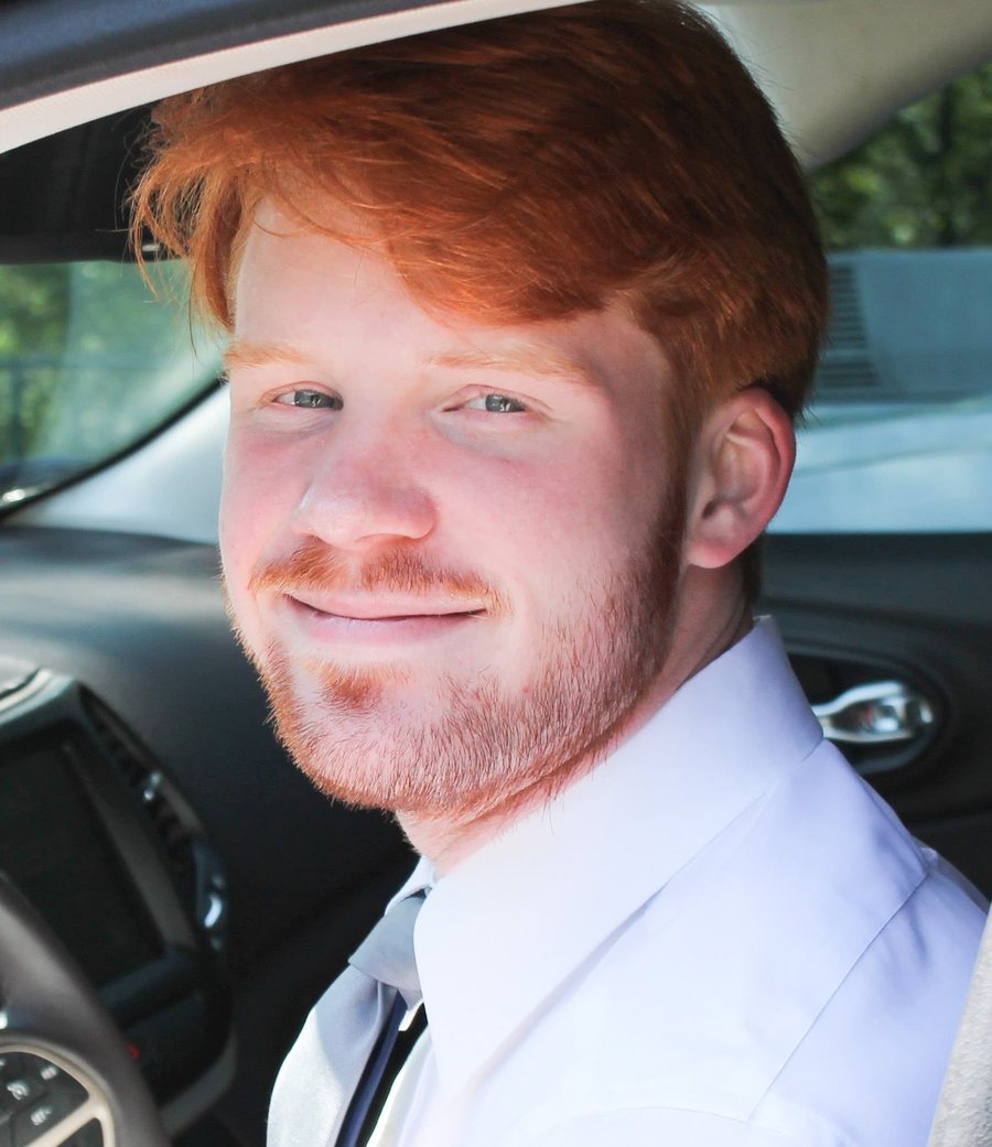 Max Cochran, smiling, sitting in the driver's seat of a car in a white shirt and tie